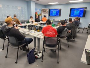 A group of people sitting around tables in front of a tv.