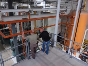 Two men are standing in a room with pipes.