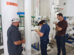 Three men standing in a room next to pipes.