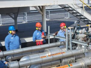Three men in hard hats are standing next to pipes.