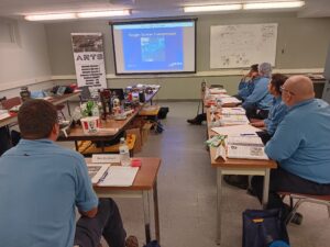 A group of people sitting at tables in front of a projector screen.